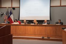 Six of the Board members seated at the dais in the meeting room. A projected image of the meeting's agenda is behind them, along with the American and Texas flags. 